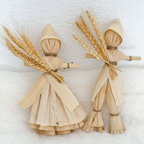 Two corn husk doll figurines with wheat on a white background